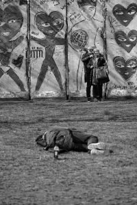 Obdachloser liegt auf Wiese vor mit Herzen bemalter Mauer, vor der ein Paar ein Selfie macht, ohne ihn zu beachten