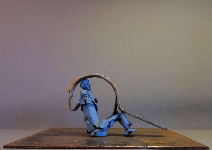 Blue metal figure sitting on a metal platform with a bent metal band going over its knees and head to its back
